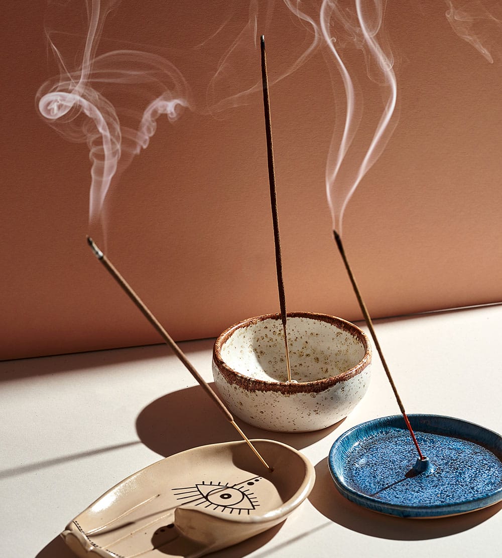 Three incense sticks in ceramic holders emit curling smoke against a warm background.