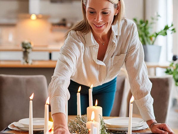 Candle sticks on a dining table are lit while a woman is leaning over them to set the table. 