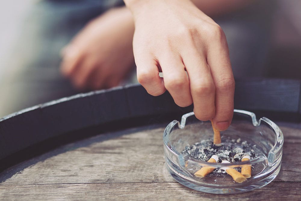 A hand stubs out a cigarette in a clear ashtray filled with cigarette butts, on a wooden surface.