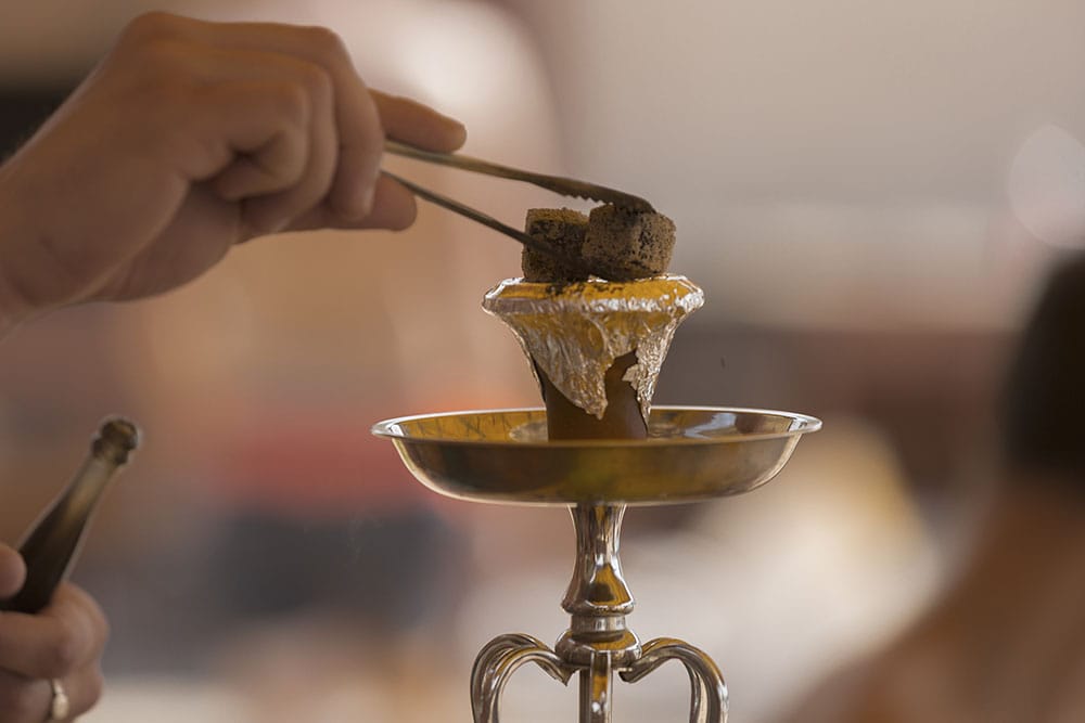 A person uses tongs to place coal on a foil-covered hookah bowl for a shisha.