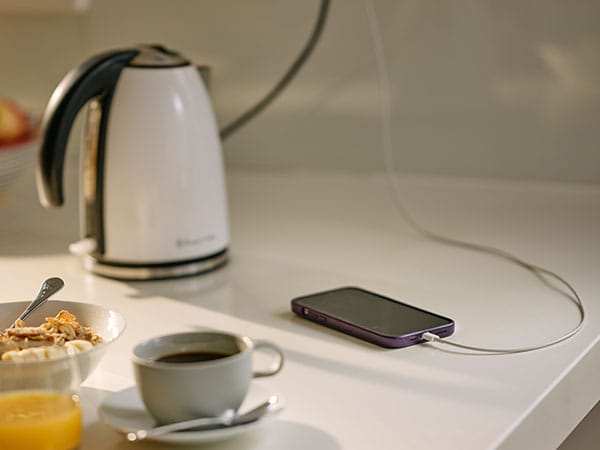 A mobile phone charging on a white stone kitchen bench top surrounded by a kettle, cup of black coffee, glass of orange juice and a bowl of cereal with banana on top. 