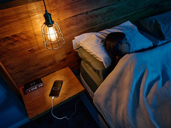 A mobile phone charging on a wooden bedside table next to an alarm clock, while a woman is sleeping in bed facing away from phone.
