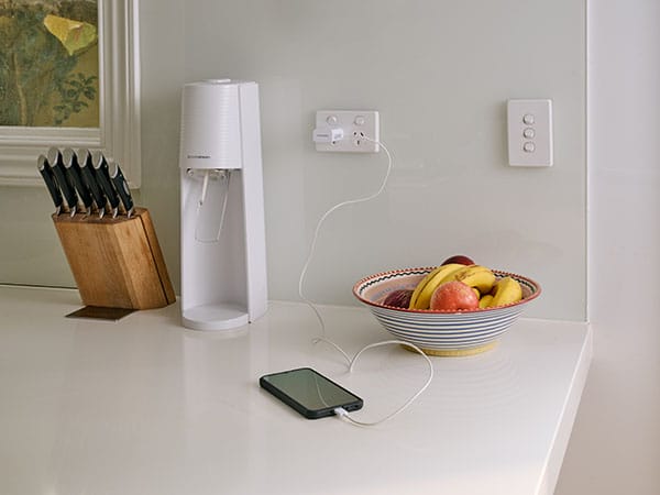 A mobile phone charging on a white cord sits on a white stone kitchen bench top. A knife block, white soda maker, and a fruit bowl filled with apples and bananas sits in the background.