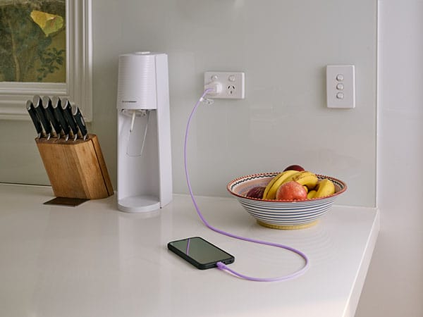 A mobile phone charging on a bright purple cord sits on a white stone kitchen bench top. A knife block, white soda maker, and a fruit bowl filled with apples and bananas sits in the background.