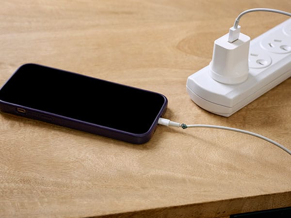A mobile phone charging with a damaged charging cord on a wooden desk . The charging cord is plugged into a white power board sitting on the corner of the desk.