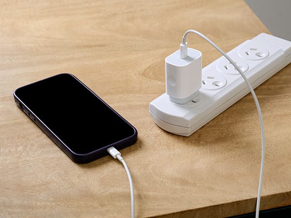 A mobile phone charging on a wooden desk. The charging cord is plugged into a white power board that is sitting on the corner of the desk.