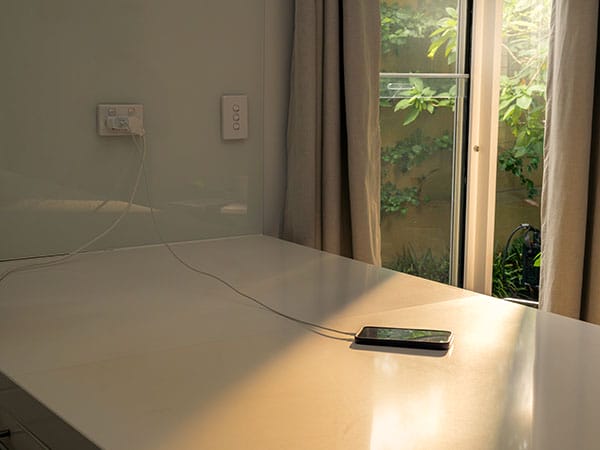 A mobile phone charging on a white stone kitchen bench top in direct sunlight. Curtains are drawn open in the background.