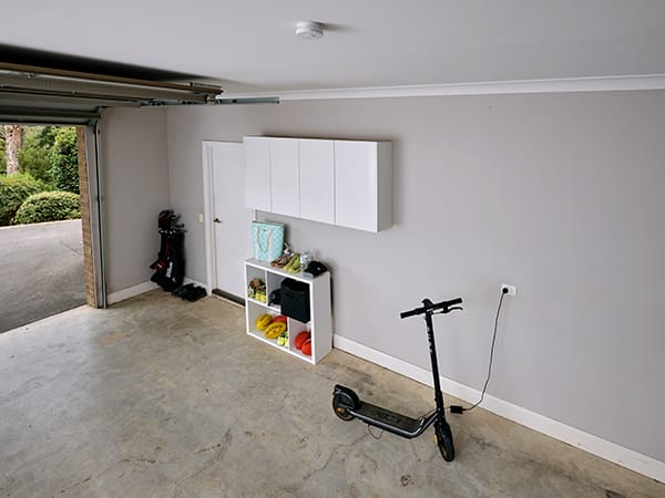 A black electric scooter charging in a garage with a smoke alarm installed on the ceiling. 