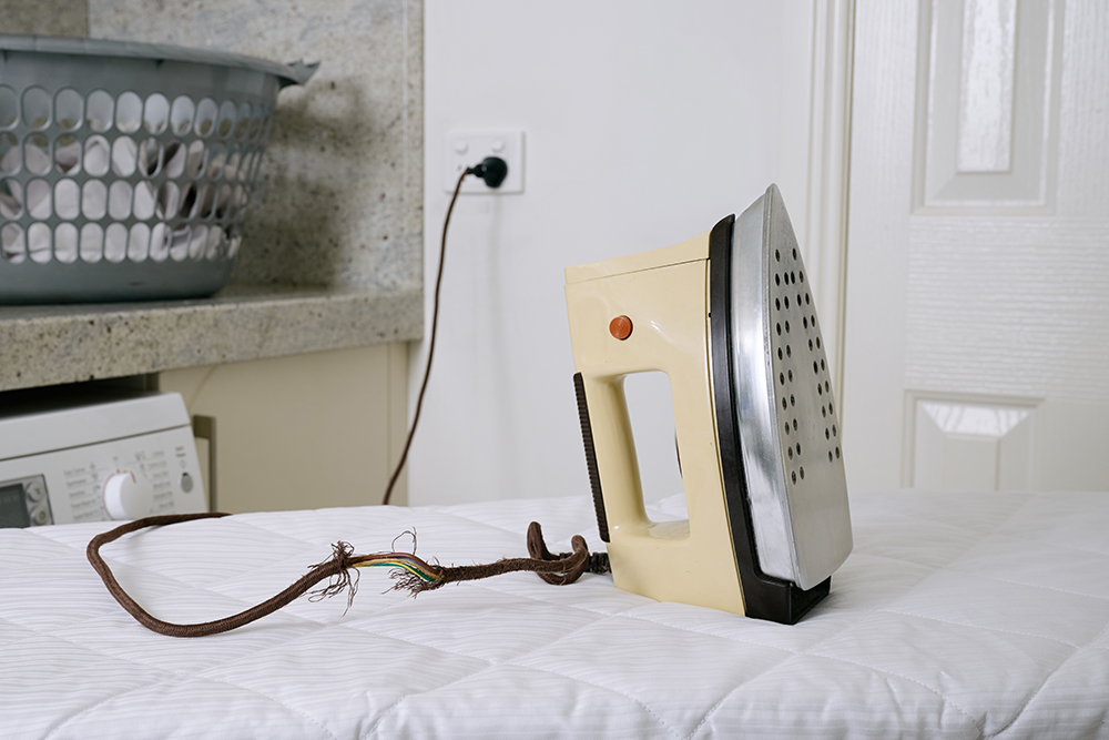 An older iron with a frayed cord sits on a padded ironing board in a laundry room. A basket of laundry and a washing machine are visible in the background.