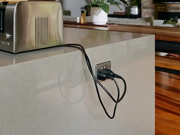 A stainless steel toaster placed on a on a kitchen stone benchtop. Its power cord dangles over the edge and is neatly plugged into power point on the end of the bench.