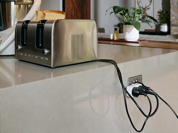 A stainless steel toaster sits beside a food processor on a kitchen stone benchtop. The power cords dangling over the edge into a double adaptor that is also plugged into a power point on the edge of the bench.