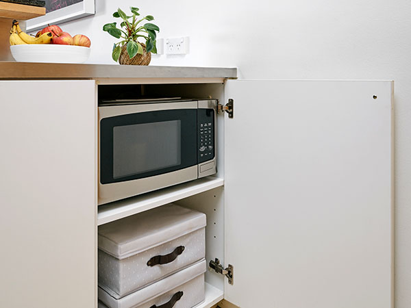 Microwave in a cupboard on top shelf. Decorative indoor plant and fruit bowl placed on bench top above. 