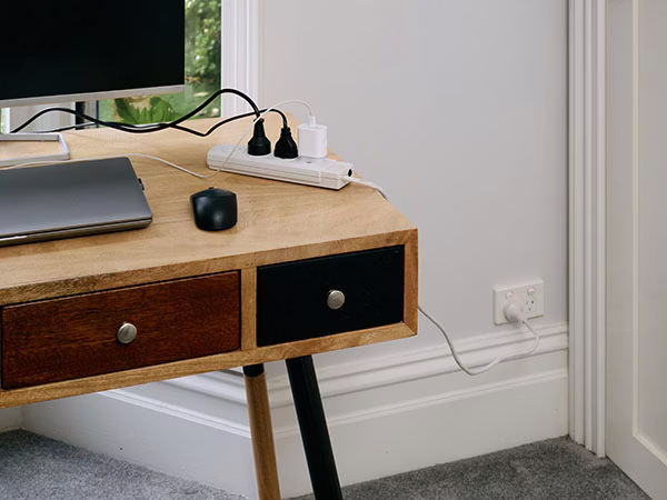 A wooden desk with laptop and monitor plugged into a power board. The power board is plugged into a power point on wall.