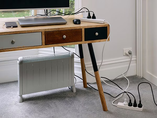 A wooden desk with laptop, monitor and mobile phone plugged into a power board. This power board is then plugged into another power board that is resting on carpet that has a heater and two other appliances plugged in. This power board is then plugged into a power point on the wall.