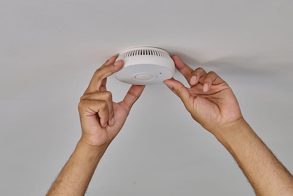 Hands installing a white smoke detector on a ceiling.