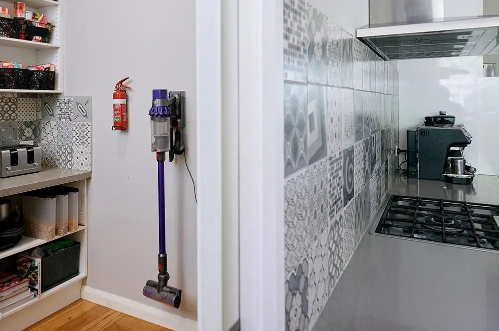 A tidy pantry with various items on the shelves, a fire extinguisher and handheld cordless vacuum stick mounted on the wall. A door way leading into an adjacent kitchen featuring a built in stove top, range hood and coffee machine. 