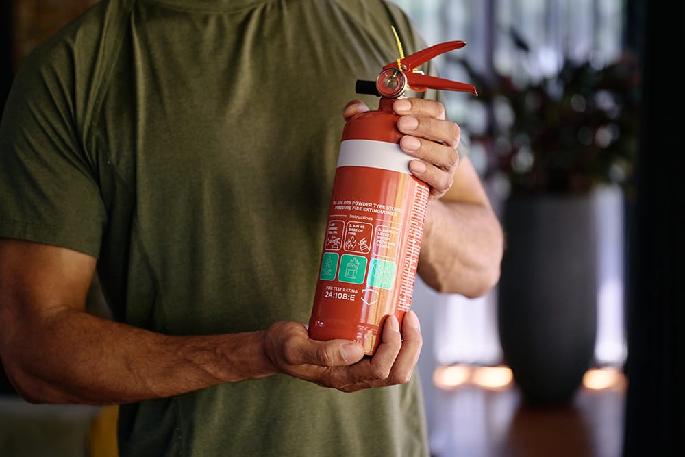 A man in his lounge room holding a red fire extinguisher with a white band. 