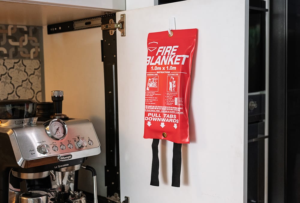 A fire blanket mounted with a plastic hook on the back of a cupboard door next to a coffee machine in a kitchen.