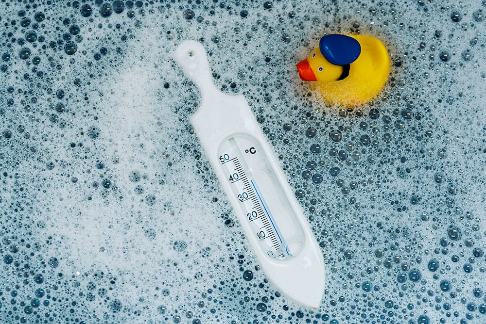 A floating baby bath thermometer and a yellow rubber duck with a blue hat in bubbly water.