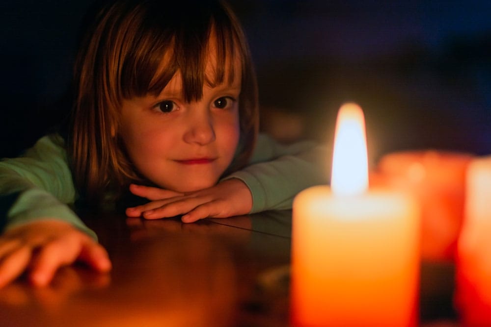 A child watching a lit candle on a table in a dark room.