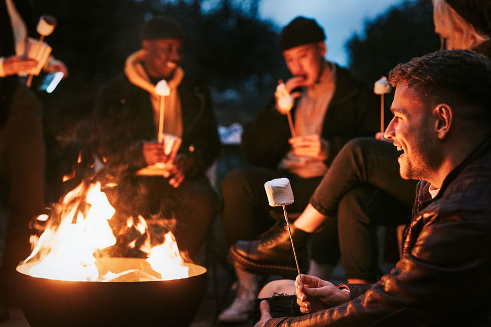 A group of friends gathered around a fire pit at dusk, smiling and roasting marshmallows on sticks.