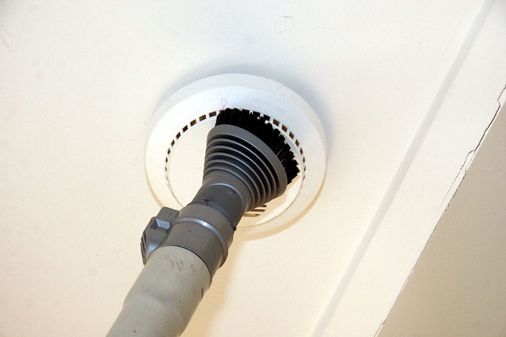 A vacuum cleaner hose with a brush attachment cleans a smoke alarm on a ceiling.