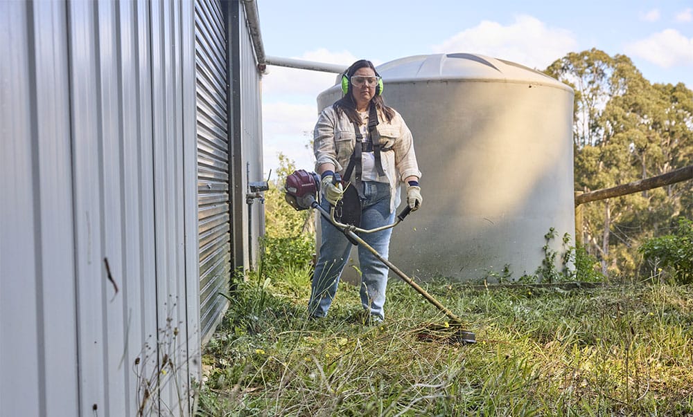 A woman is using a petrol powered week trimmer to shorten grass behind a shed. A water tank and gum tree forest is in the background.