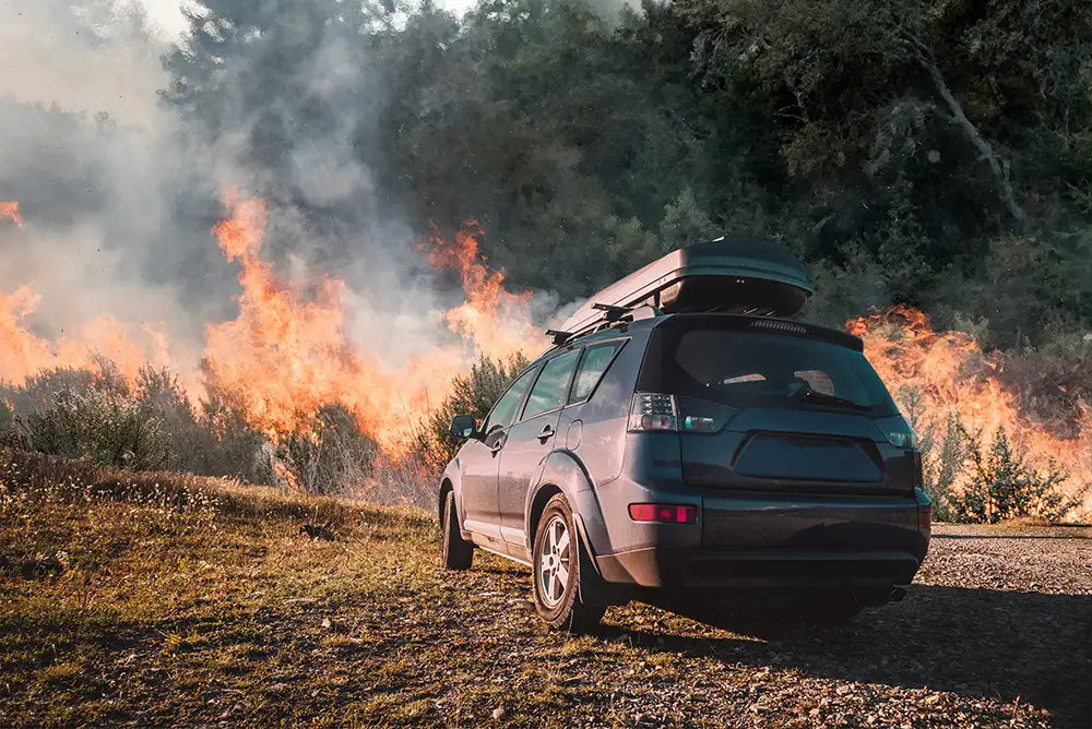 A car parked in a clear space while a fire burns in scrub nearby.