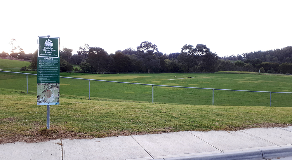 A sign that says 'Bushfire Place of Last Resort' in front of a grass covered sports oval, ringed by trees.
