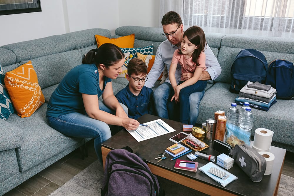 A family sitting together in a living room, discussing an emergency plan document with supplies such as water, canned food, flashlight, and a first aid kit laid out on the coffee table.
