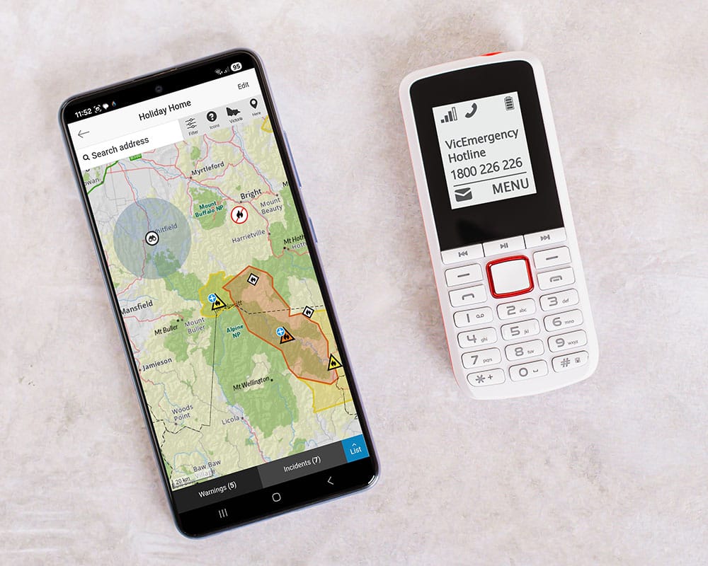 Two phones. On the left is a modern smart phone with the VicEmergency app displaying a map of part of Victoria indicating fires in the area and yellow and orange Advice and Watch and Act alerts. It also indicates that it is a Total Fire Ban day. On the right is an older style push button phone displaying the VicEmergency Hotline number 1800 226 226.