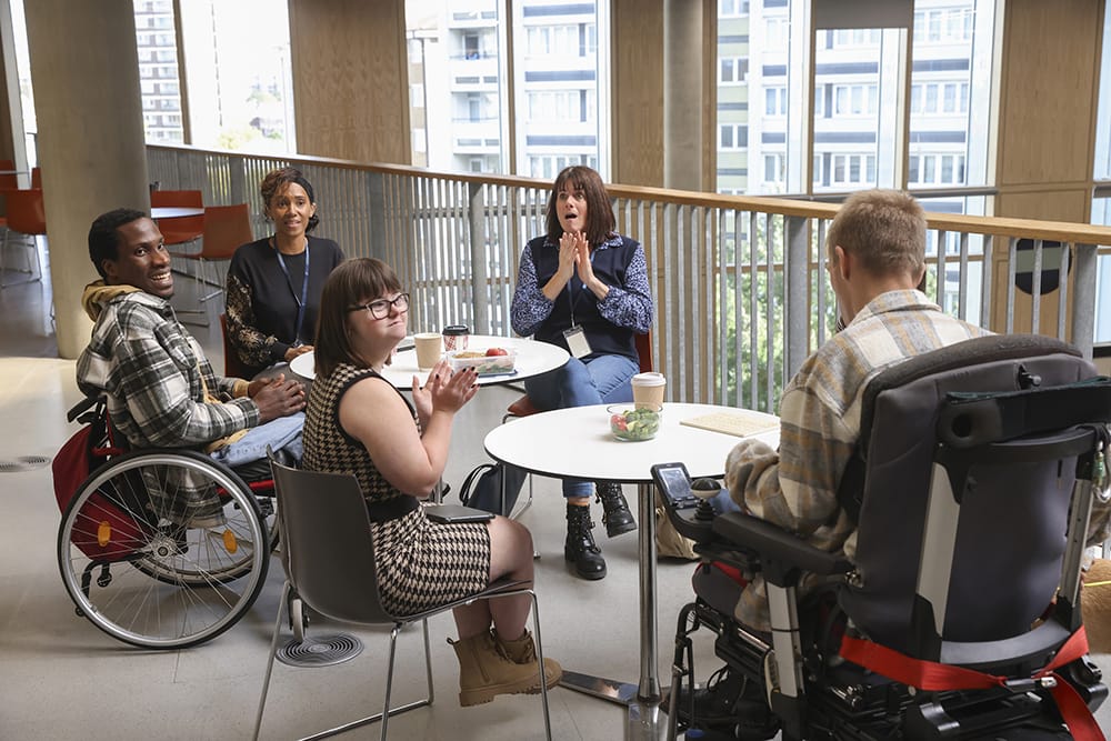 A group of people with various disabilities, some in wheelchairs or using sign language, chatting around two circular table in a modern indoor space.