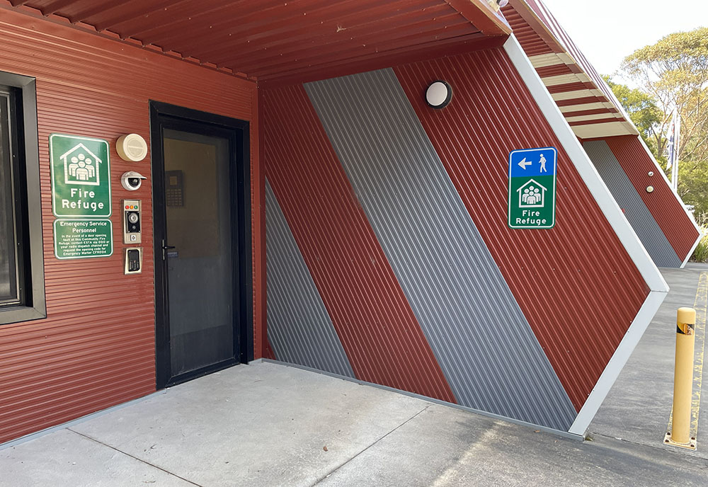 Entrance to a modern looking building. There are signs that say Fire Refuge, a security camera, buttons with red, yellow and green lights, a speaker and a keypad.