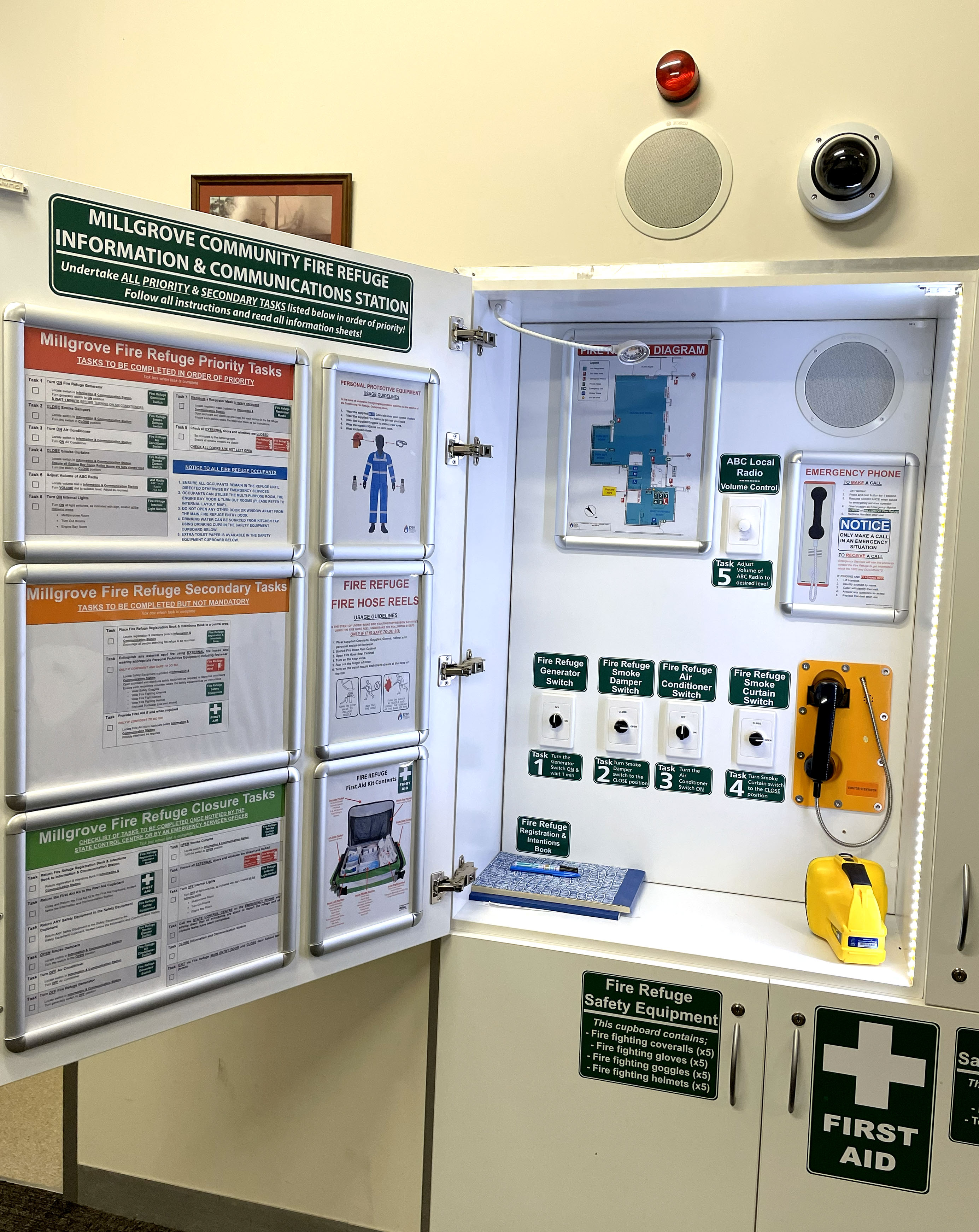 An open cabinet with many notices on the inside of the door providing instructions. Inside the cabinet is a floorplan, five numbered switches and a telephone handset. There is also a book, pen and torch. Beneath the cabinet is a closed cupboard with signs indicating first aid and fire refuge safety equipment.