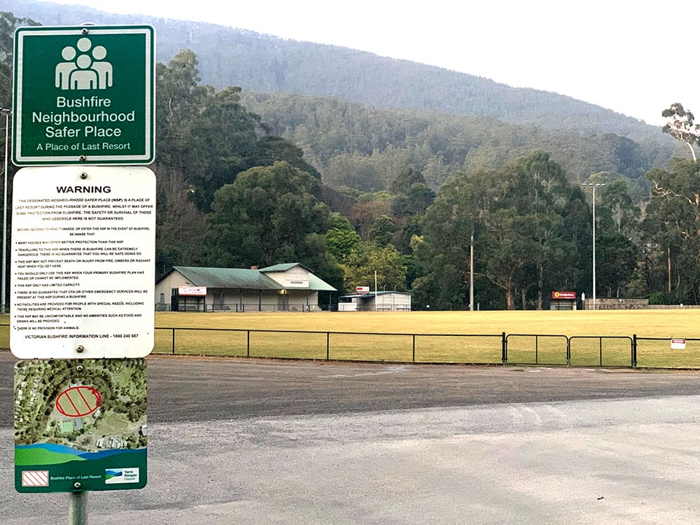 A picture of a fenced sportsfield. The oval is ringed with trees and there is a tree-covered mountain behind the oval. In the foreground is a sign attached to a pole which reads: 'Bushfire Neighbourhood Safer Place - A Place of Last Resort'.