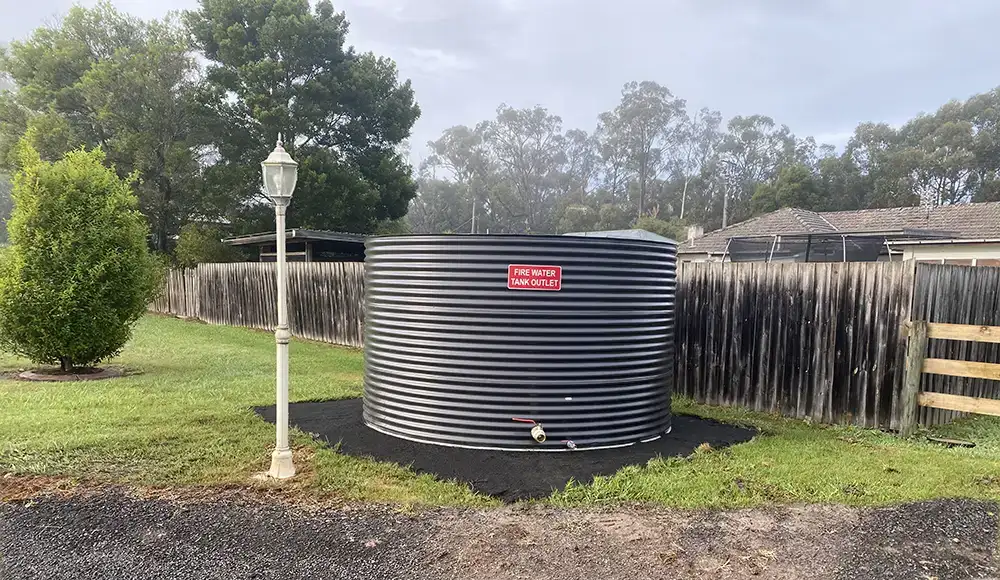 A steel water tank near a backyard fence with Fire Water Tank Outlet written on it. The tank has a large firefighting outlet. It sits next to a gravel driveway and a lamp post is nearby.