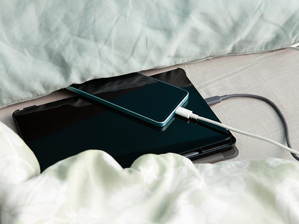 A mobile phone and tablet on a bed resting between soft pillows and a blanket while unsafely charging. 