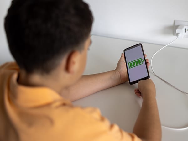 A young boy in a yellow shirt sitting at a desk, holds a mobile phone connected to a charger. The phone displaying a full battery icon.
