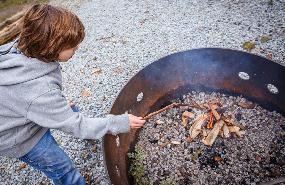 A young boy in a gray hoodie and jeans tends a small fire in a metal fire pit with a stick. The ground is covered with gravel.
