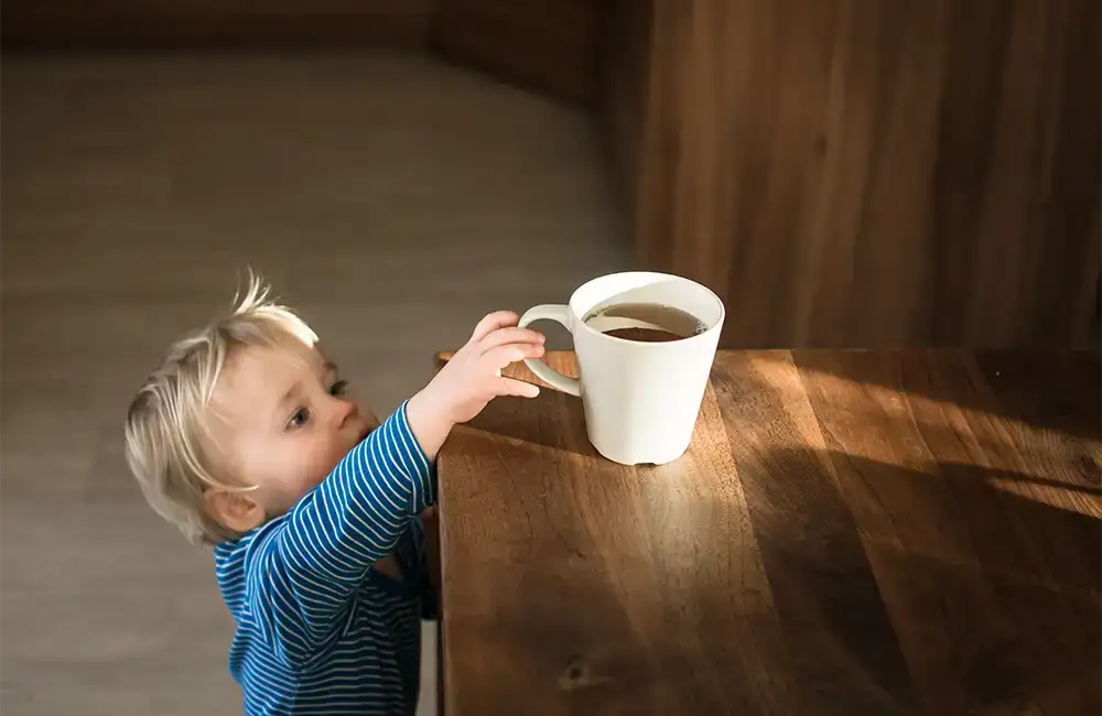 A toddler reaching up to grab a hot cup of coffee from a high table.