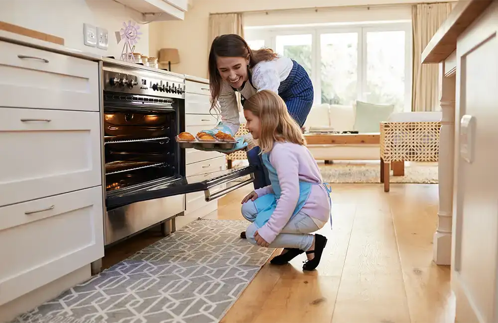 A woman in a modern kitchen removing muffins from an open even while a young girl watches and learns.