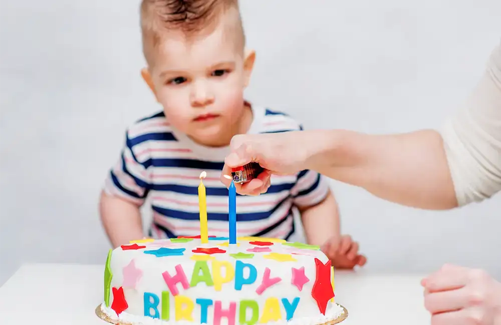 Young boy in striped shirt looks at blue candle being lit on a cake that says happy birthday