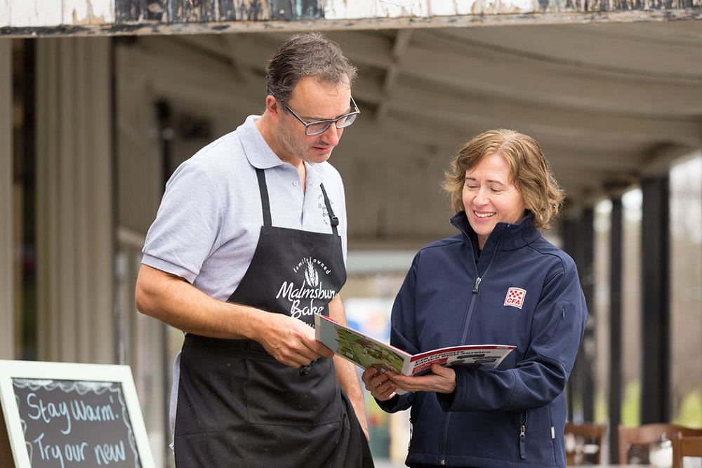 A man in a bakery apron and a woman in CFA uniform stand outside a bakery, looking over a fire safety information booklet.