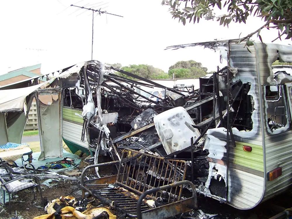 Charred remains of a burnt-out caravan, with twisted metal and melted parts, sit in a grassy area, conveying destruction and loss.