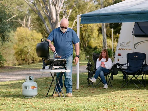 Man cooking on a portable barbecue next to a caravan awning on a grassy campsite. A woman sits nearby in a camp chair, holding a drink.