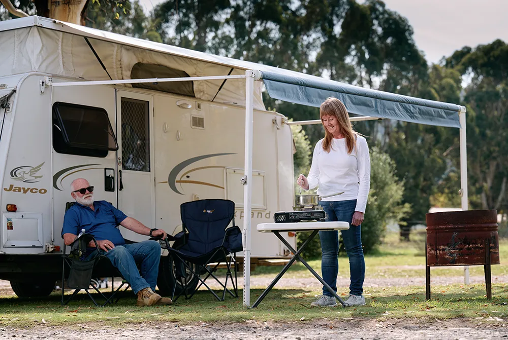A man relaxes in a camping chair under a caravan awning, while a woman cooks at a portable stove. The setting is peaceful, surrounded by trees.