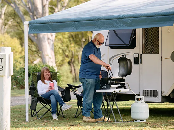 Man cooking on a portable barbecue under a caravan awning on a grassy campsite. A woman sits nearby in a camp chair, holding a drink.