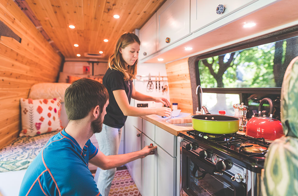 Caravan interior with wooden walls features a young woman at a sink and a man opening a cabinet. An oven and stove top with pots is visible.