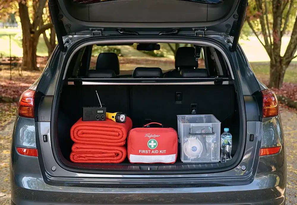 Dark grey car parked on a gravel driveway with the tailgate open. Inside contains emergency supplies including red woolen blankets, a first aid kit, yellow flashlight, portable radio, container with P2 masks and water bottles.