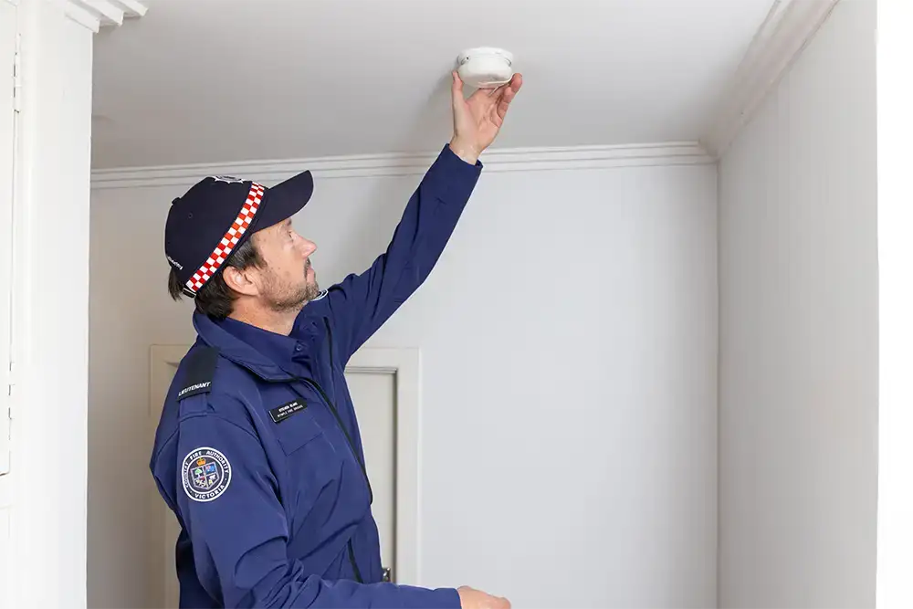 A man in a CFA uniform installs a smoke alarm on the ceiling of a hallway